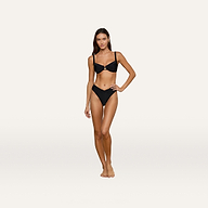 A woman stands barefoot on a white background, wearing the Adrienne Bikini Top in black—a balconette bikini top with subtle trapunto stitching and underwire support. She has one hand on her hip and looks forward with a neutral expression.