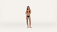 A woman stands barefoot against a plain white background, wearing a black bikini. She faces forward with one arm crossed over her stomach and the other arm resting on it.
