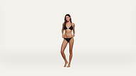 A woman stands barefoot against a plain white background, wearing a black bikini. She is posing with her arms slightly bent and looking toward the camera.