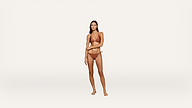 A woman stands on a plain white background wearing a brown bikini, looking at the camera with a neutral expression, her right arm crossing her body and her left arm touching her right elbow.