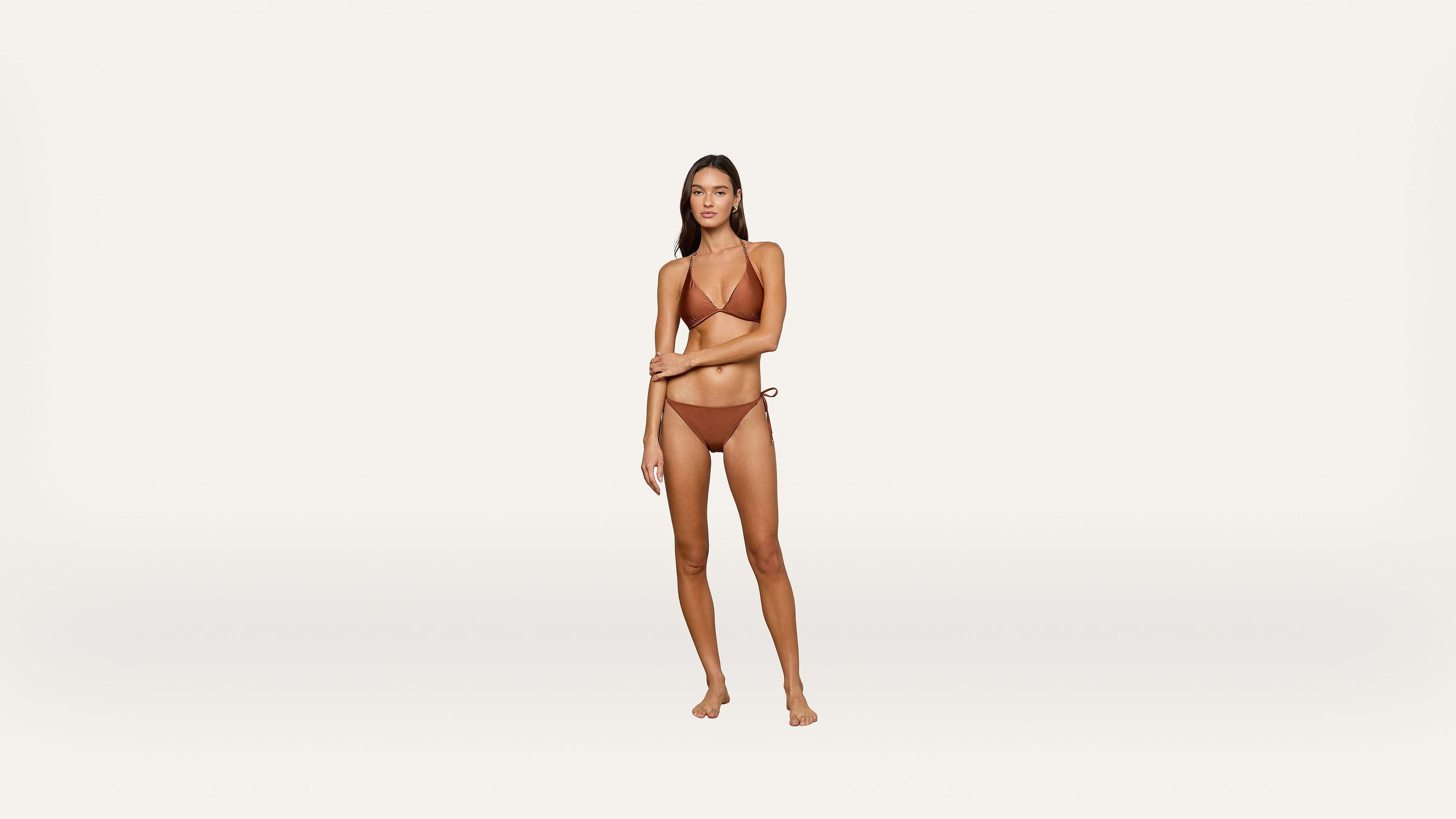 A woman stands on a plain white background wearing a brown bikini, looking at the camera with a neutral expression, her right arm crossing her body and her left arm touching her right elbow.
