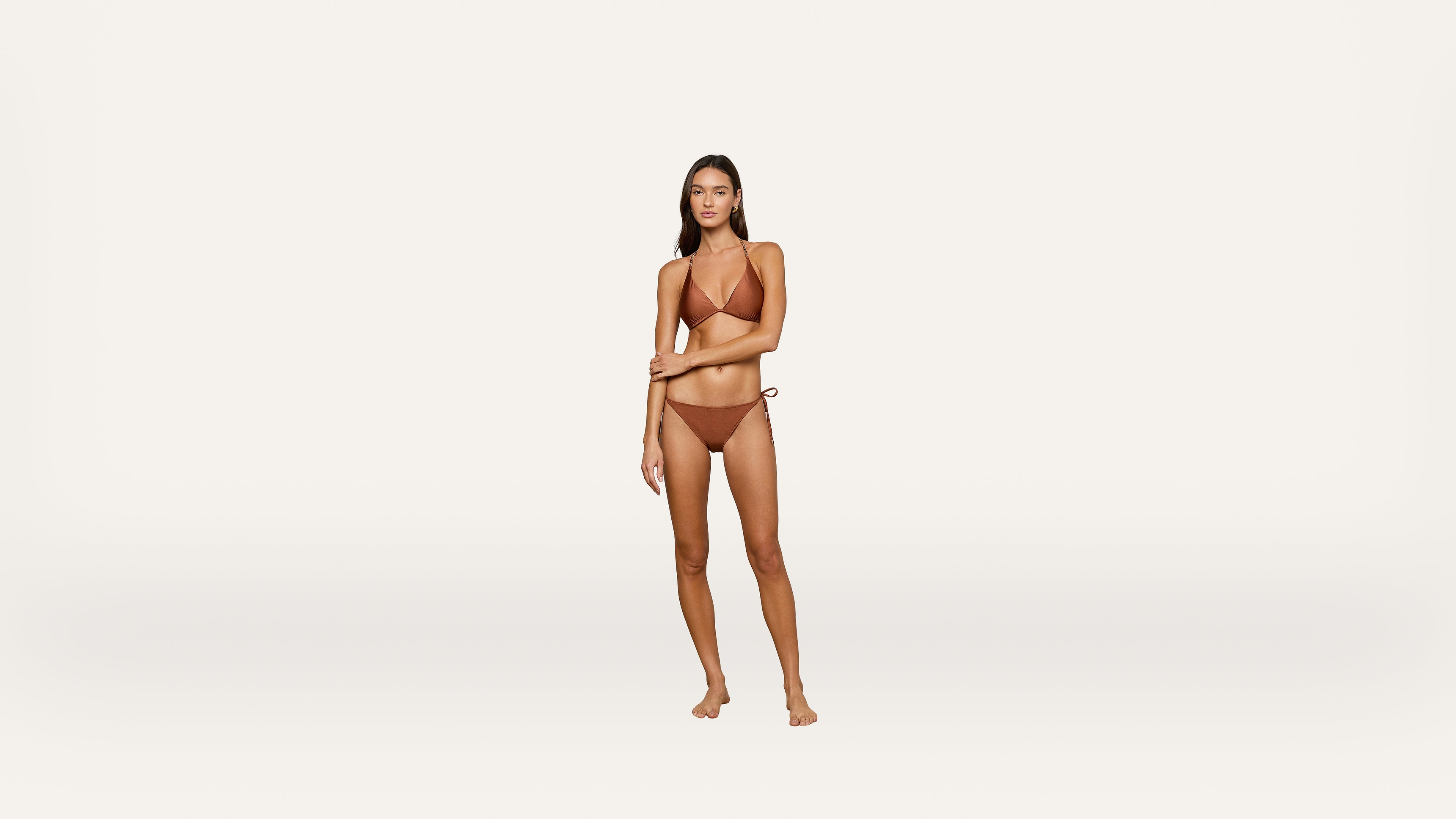 A woman stands on a plain white background wearing a brown bikini, looking at the camera with a neutral expression, her right arm crossing her body and her left arm touching her right elbow.