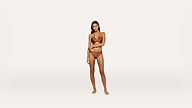 A woman stands barefoot against a plain white background, wearing a brown bikini. She faces forward with one hand resting on her opposite arm.
