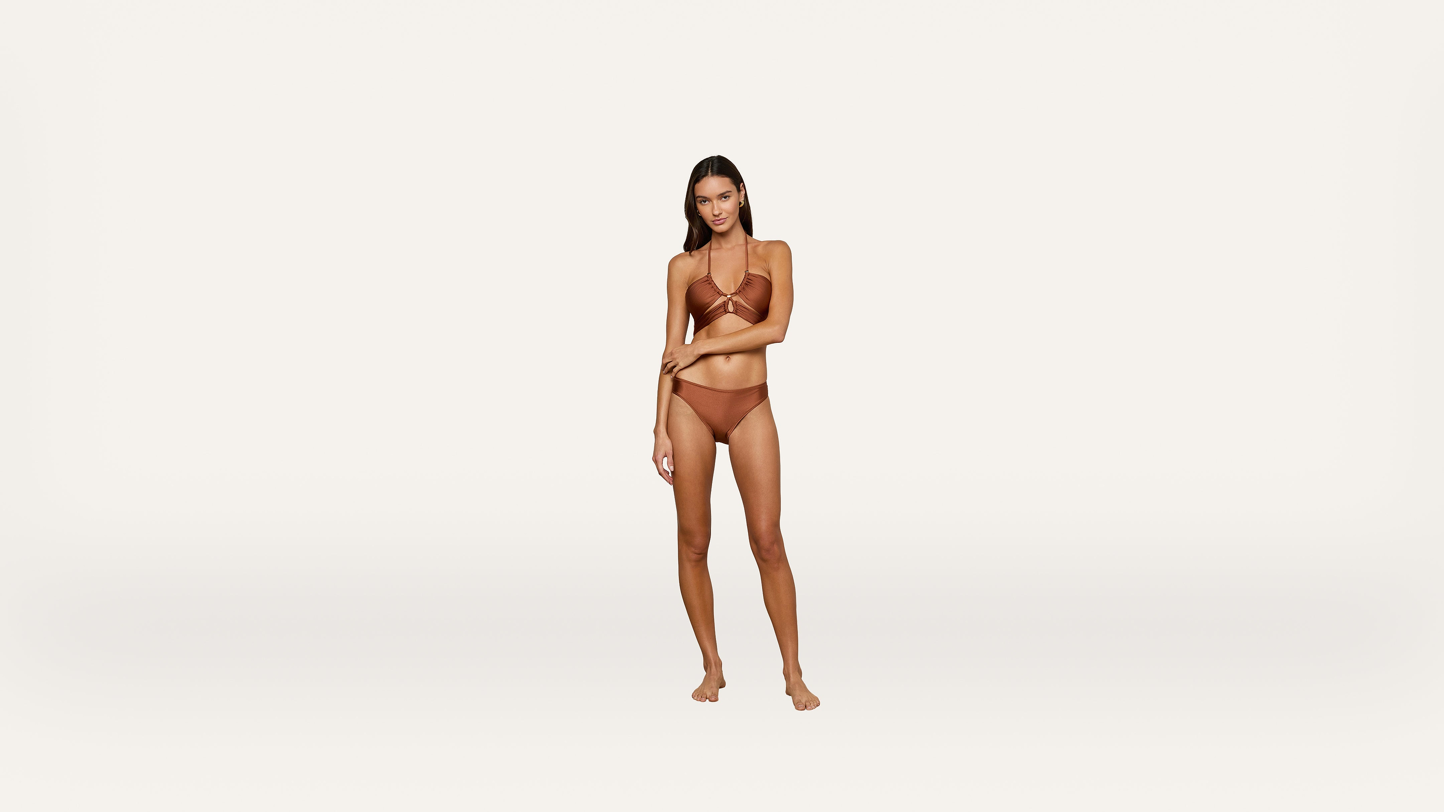 A woman stands barefoot against a plain white background, wearing a brown bikini. She faces forward with one hand resting on her opposite arm.
