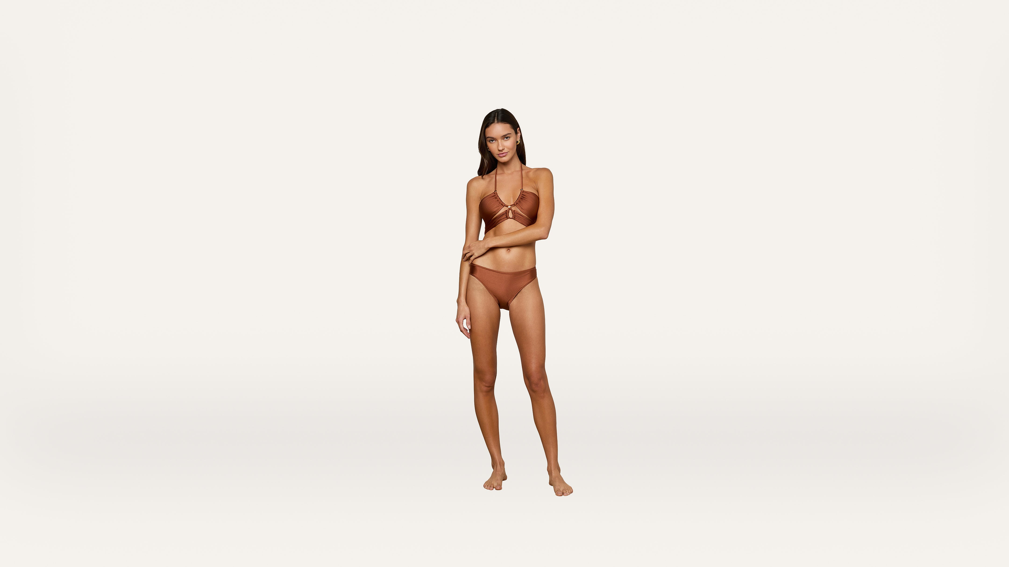 A woman stands barefoot against a plain white background, wearing a brown bikini. She faces forward with one hand resting on her opposite arm.