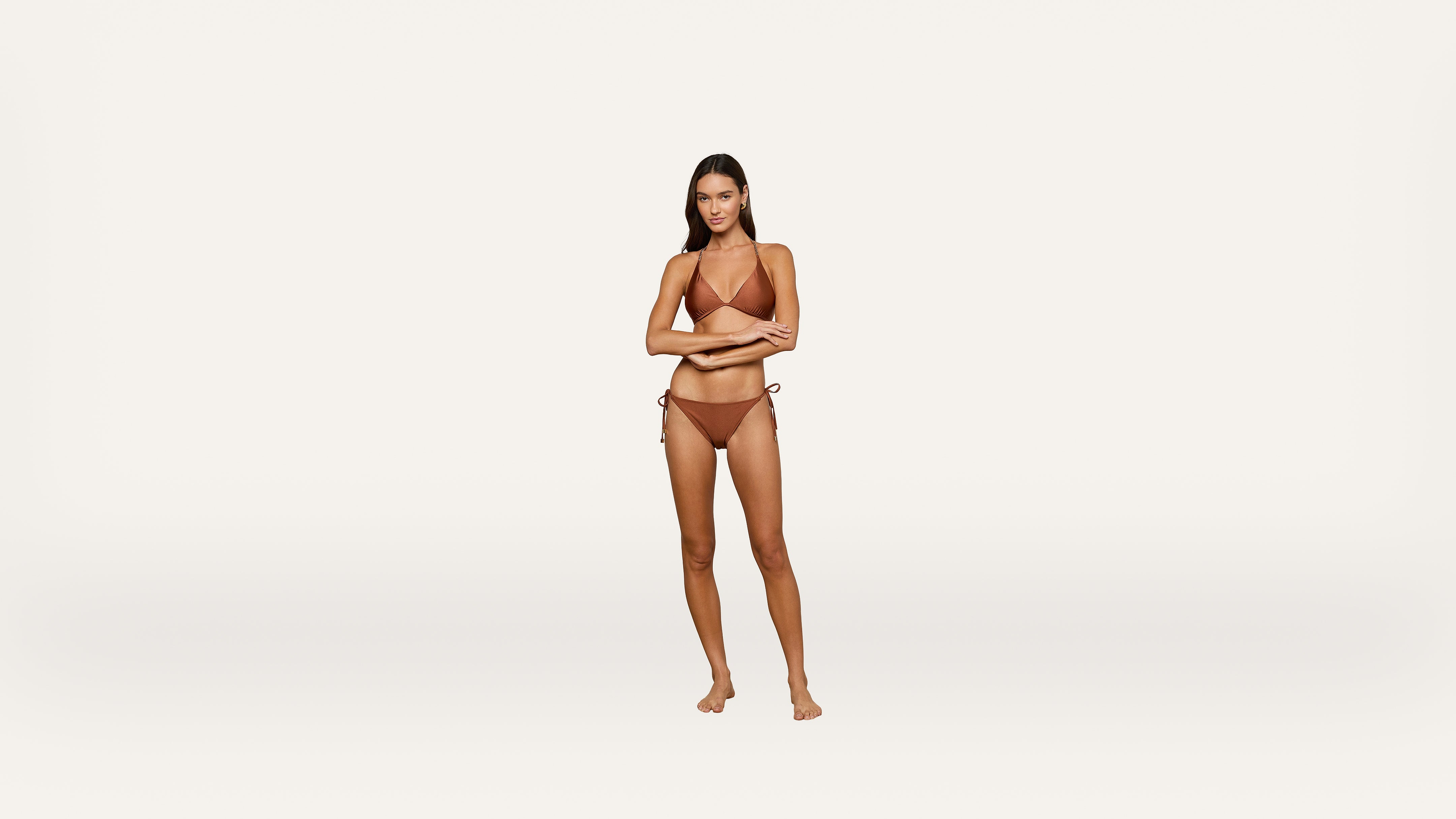 A woman stands barefoot on a plain white background, wearing a brown bikini. She has long dark hair and her arms are crossed in front of her body.