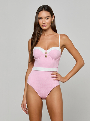 A woman poses in the Amie Bandeau One-Piece Swimsuit—light pink bouclé nylon with UPF 50, white trim, two front buttons, and thin shoulder straps—standing against a plain gray background.