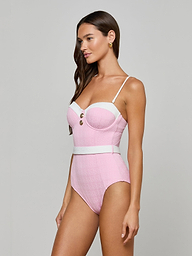 A woman models the Amie Bandeau One-Piece Swimsuit, featuring pink and white bouclé nylon, thin straps, gold buttons, and a belted waist against a plain light gray background.
