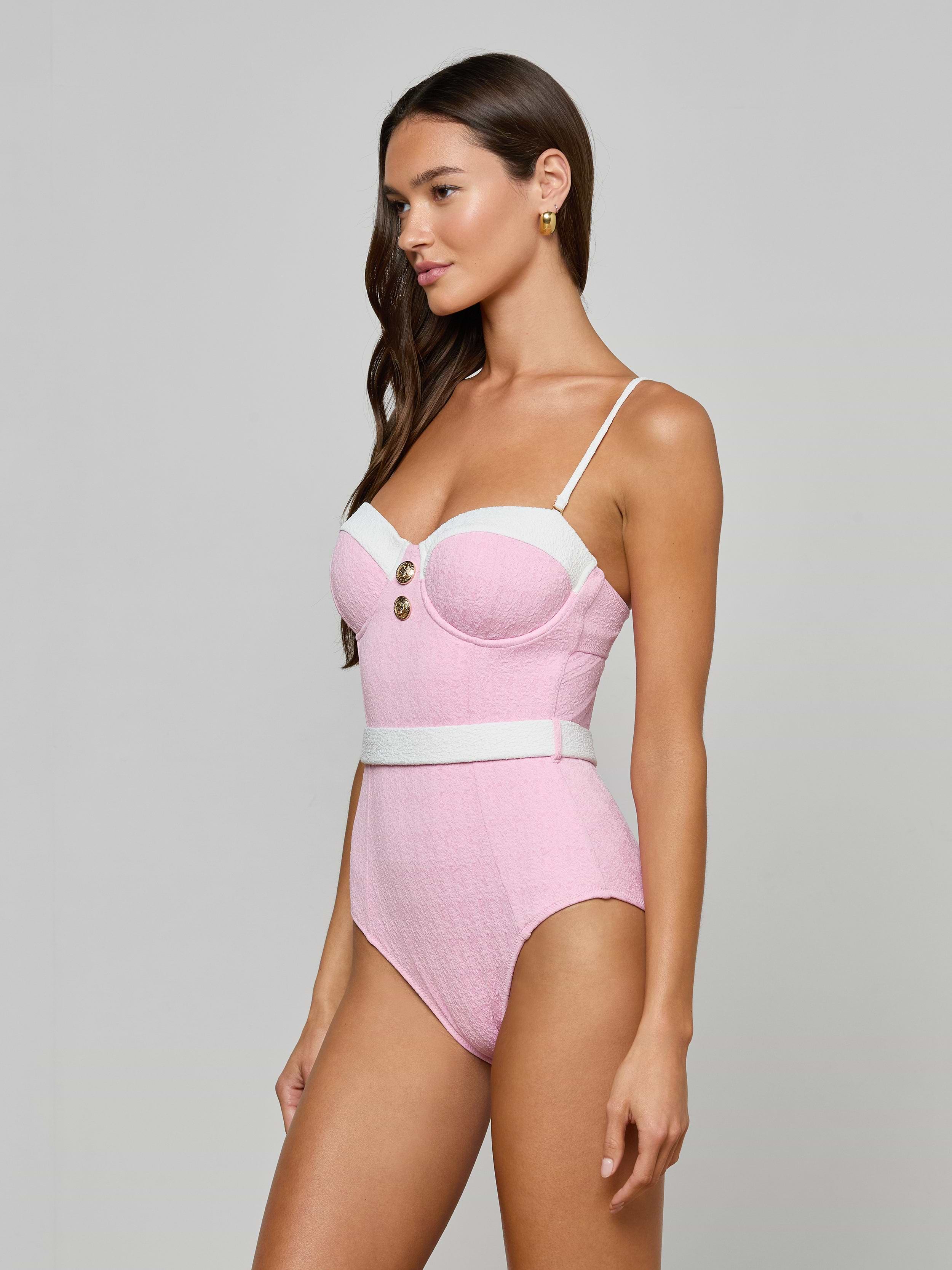 A woman models the Amie Bandeau One-Piece Swimsuit, featuring pink and white bouclé nylon, thin straps, gold buttons, and a belted waist against a plain light gray background.