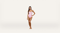 A woman stands barefoot wearing a light pink one-piece swimsuit with spaghetti straps, posing confidently against a plain, white background.