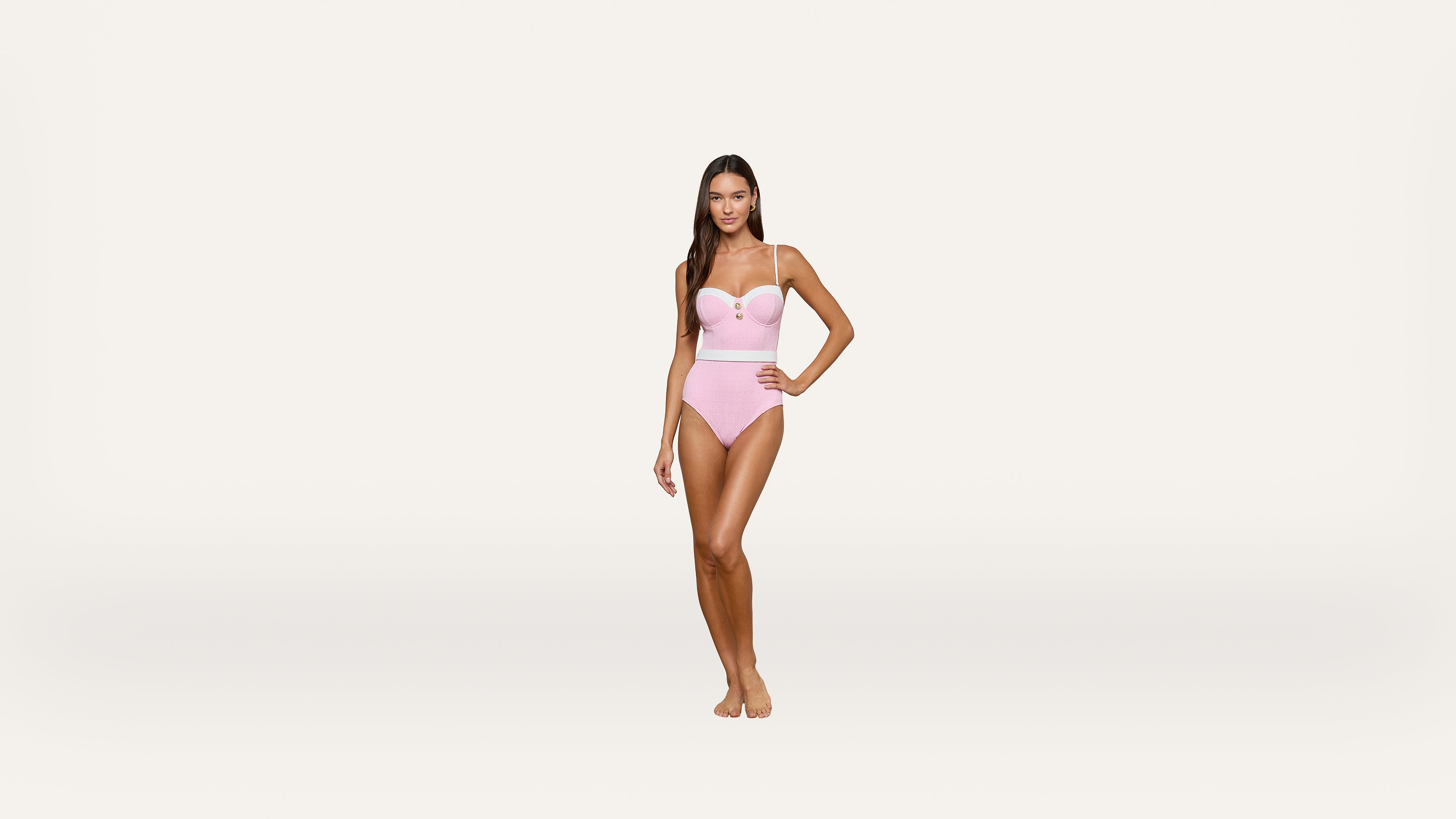 A woman stands barefoot wearing a light pink one-piece swimsuit with spaghetti straps, posing confidently against a plain, white background.