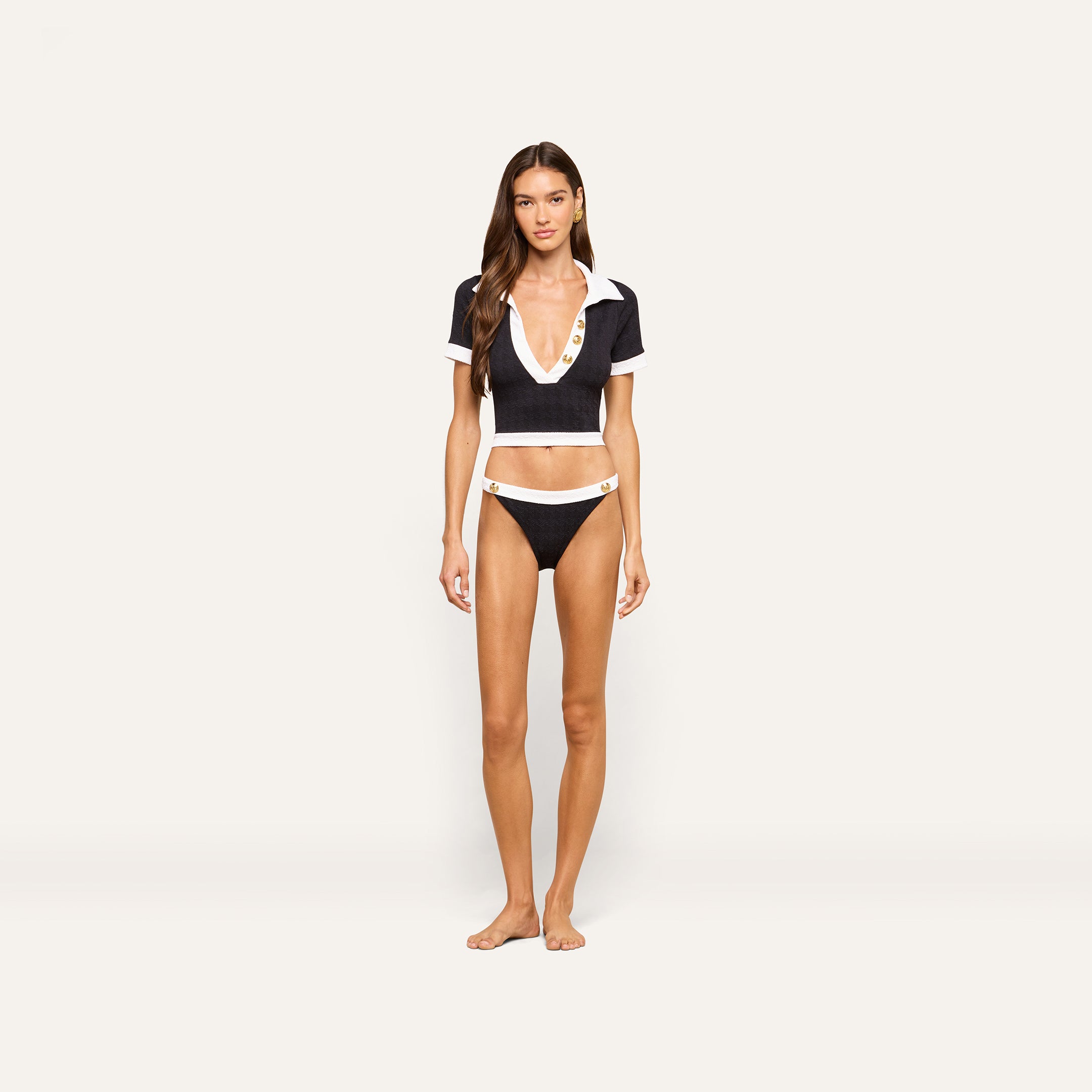 A woman stands barefoot against a plain, light background wearing a deep V-neck black and white short-sleeve cropped top paired with the Jean Bikini Bottom—matching high-leg cut bikini bottoms crafted from textured bouclé fabric.