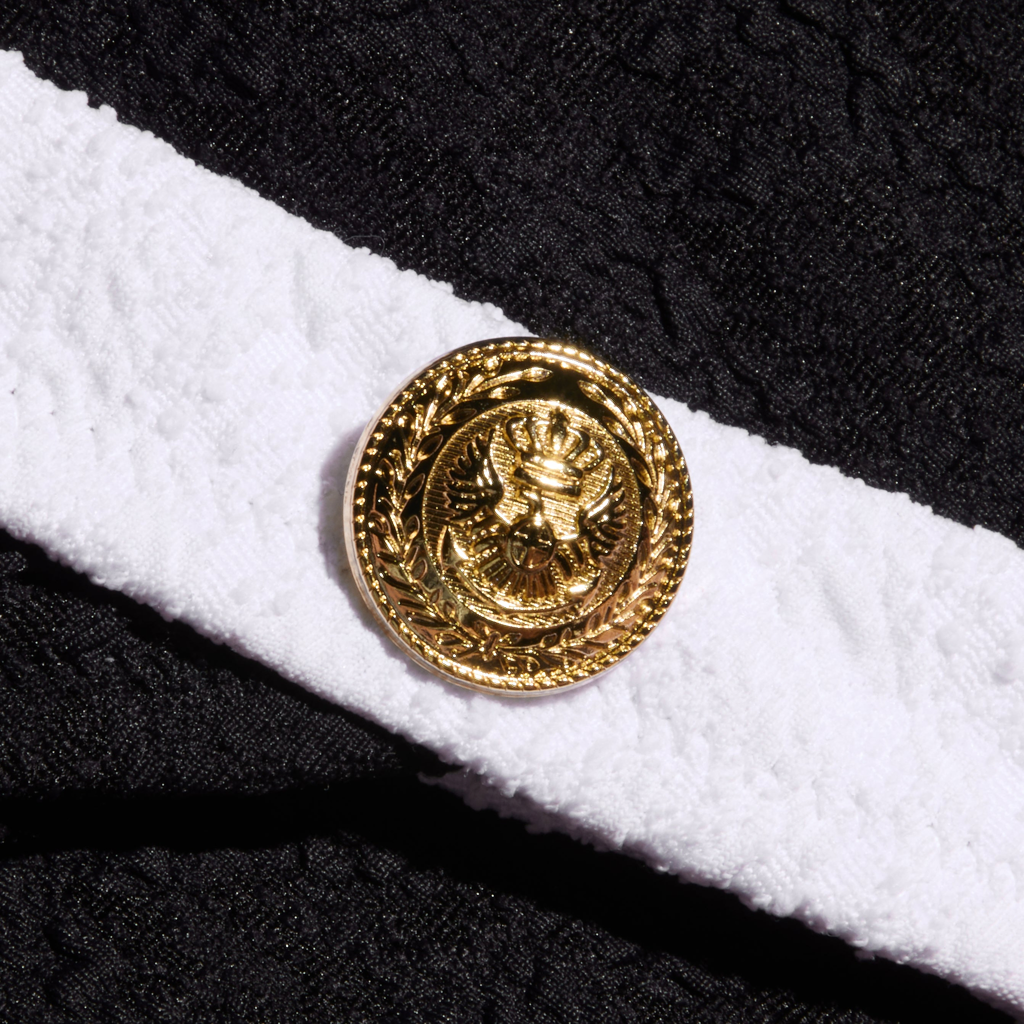 A gold button with an embossed eagle emblem is attached to a textured bouclé white band on the Jean Bikini Bottom, set against a background of black fabric.