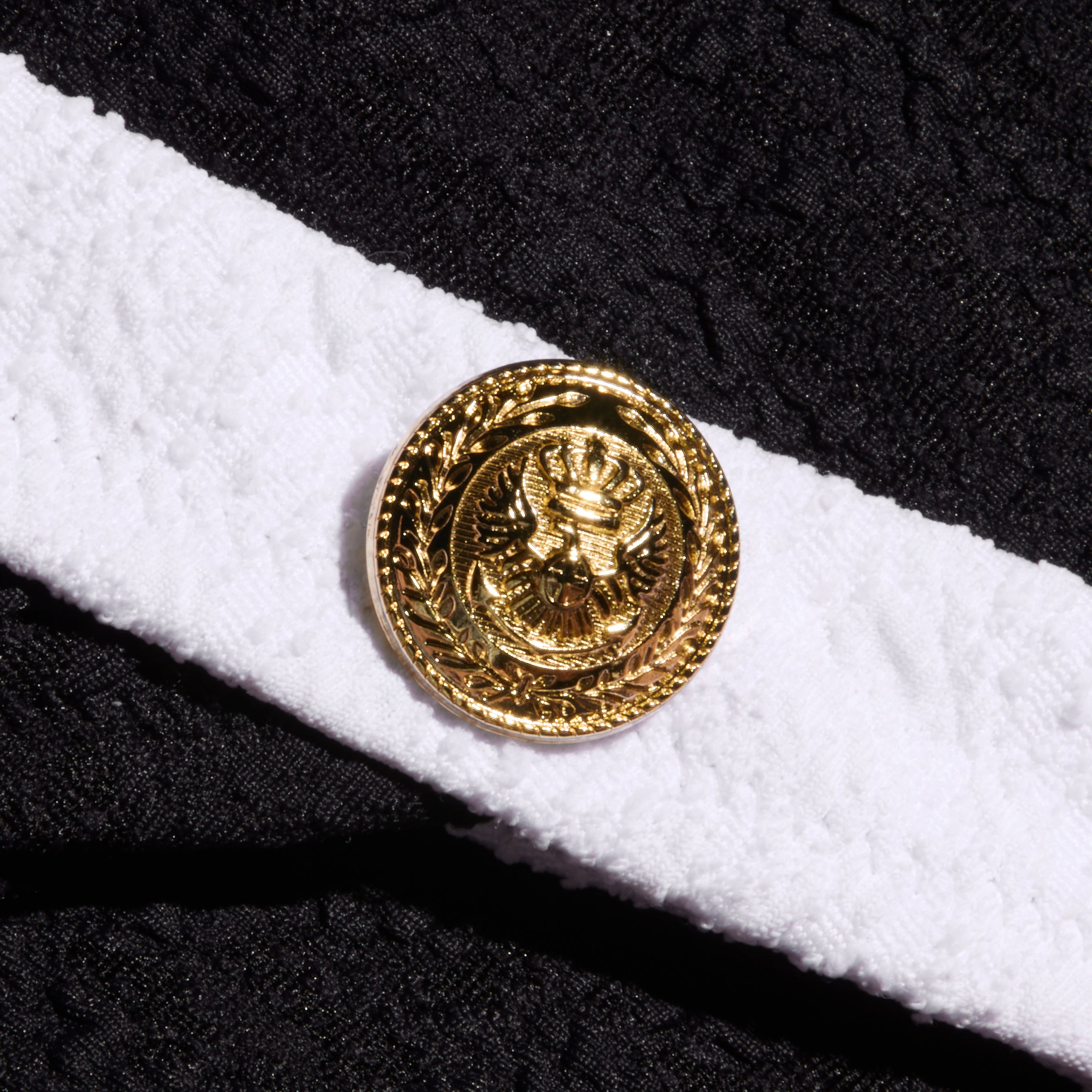 A gold button with an embossed eagle emblem is attached to a textured bouclé white band on the Jean Bikini Bottom, set against a background of black fabric.