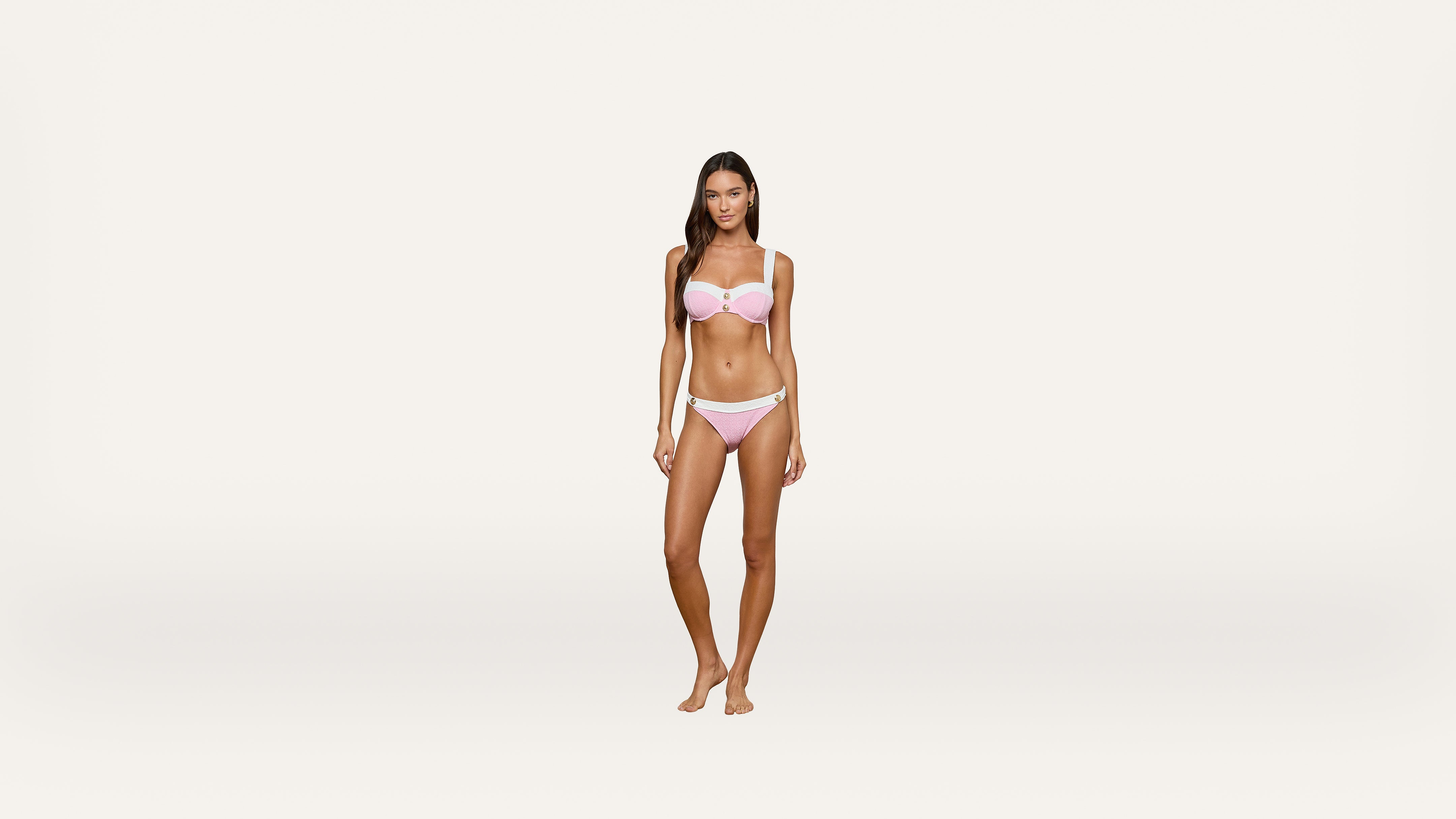 A woman standing barefoot on a light background, wearing a pink bikini and looking at the camera with a neutral expression.