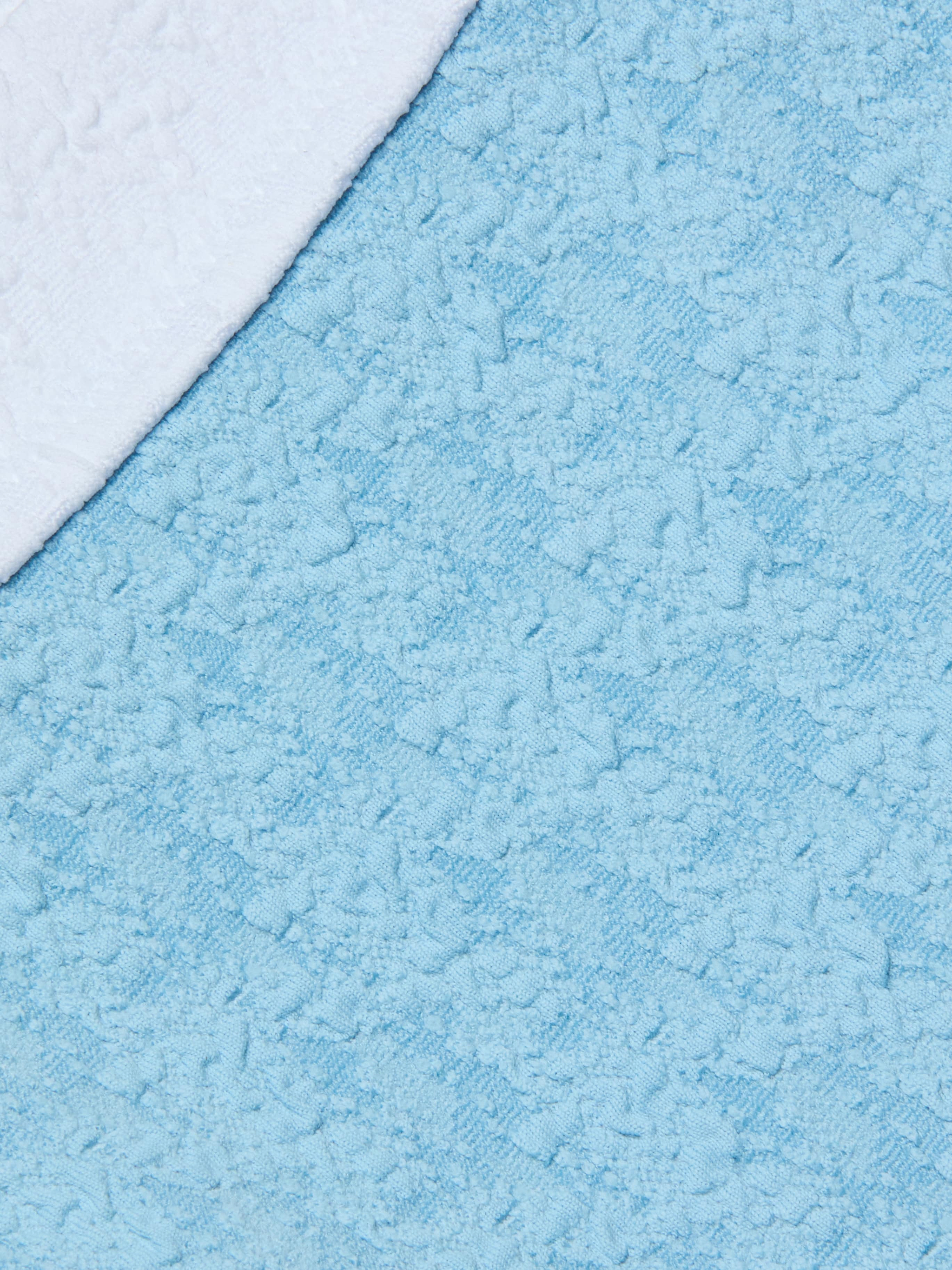A close-up of the Jean Bikini Bottom in a textured light blue bouclé fabric, with a white towel overlapping it diagonally in the upper left corner.