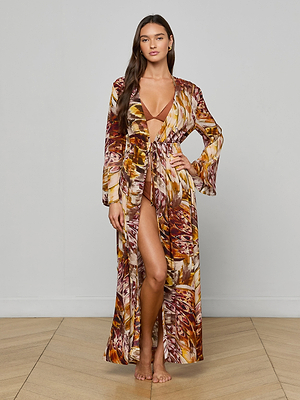 A woman stands barefoot indoors, posing against a plain light gray wall and wooden floor, wearing the Janus Cover-Up Robe with a brown, yellow, and purple abstract pattern over a matching bikini.