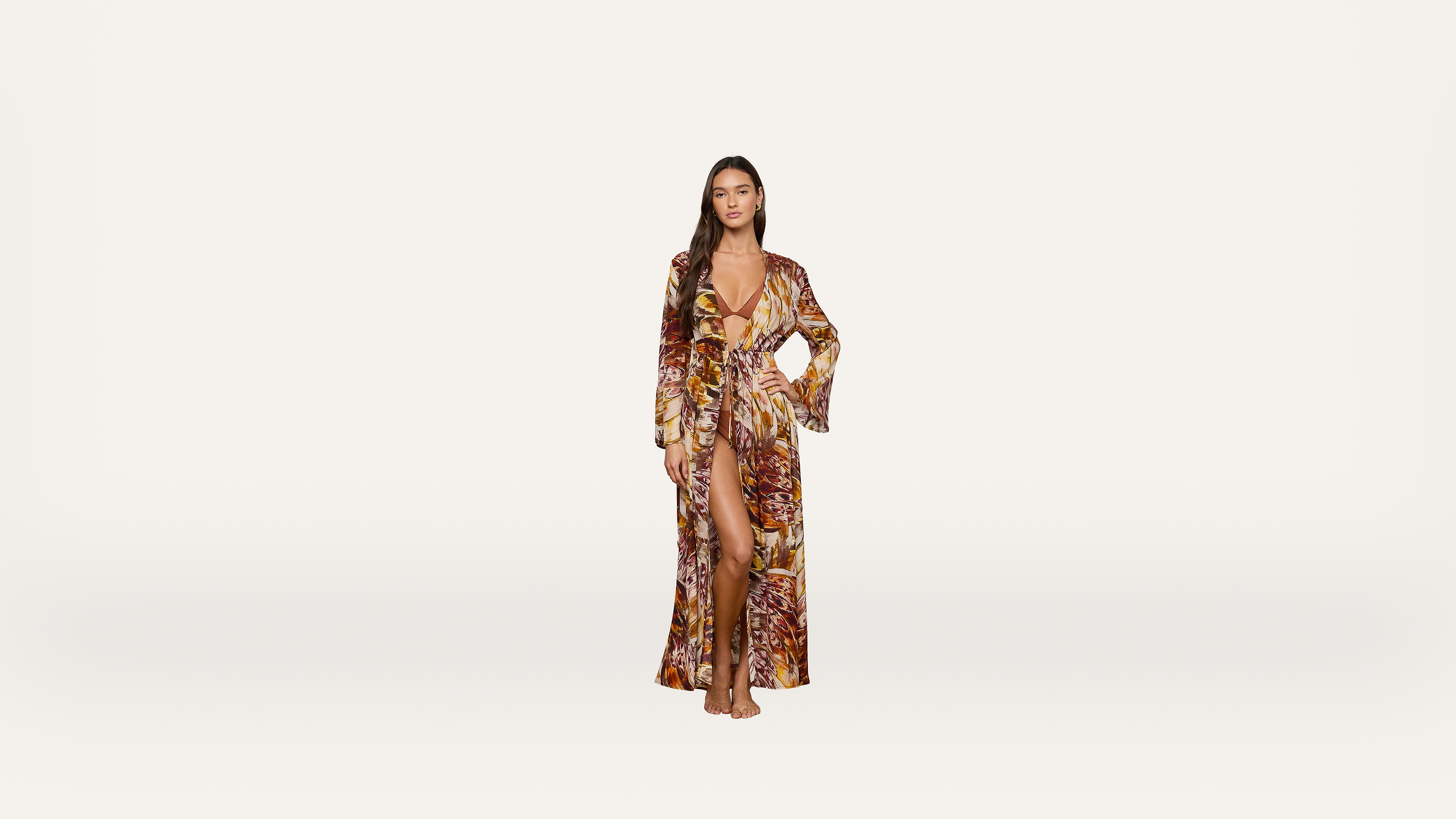 A woman stands barefoot wearing a long, patterned dress with a deep V-neck and thigh-high slit against a plain white background.