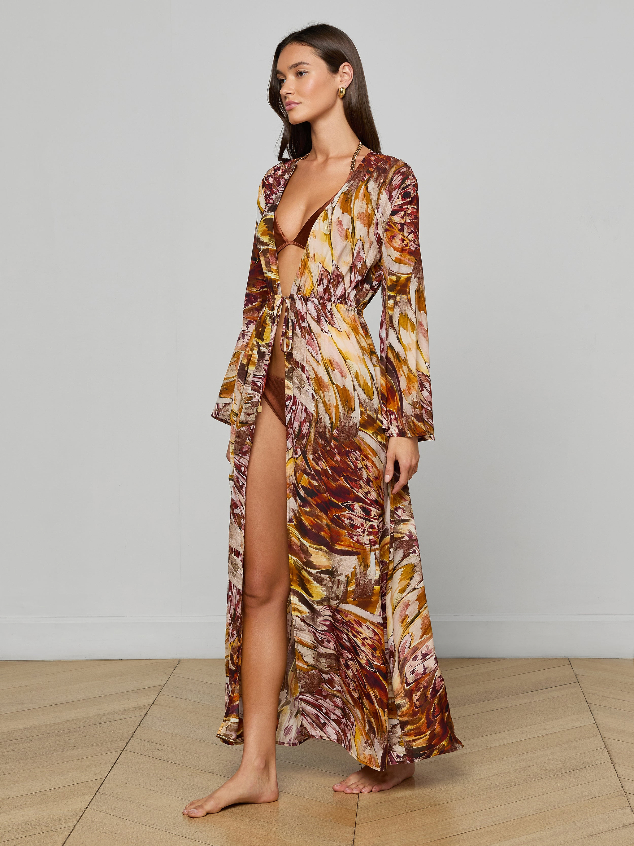 A woman stands barefoot against a plain white background wearing the Janus Cover-Up Robe, a long, flowy viscose chiffon dress with a colorful brown, orange, and yellow abstract pattern. The robe features a deep V-neck and a high slit.