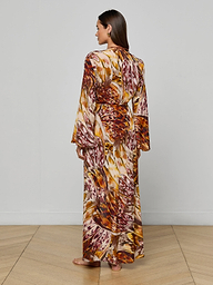 A woman stands barefoot, facing away, wearing the Janus Cover-Up Robe—a long, flowy viscose chiffon dress featuring a colorful abstract feather print in gold, orange, and brown tones—set against a plain white background.