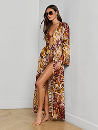 A woman poses barefoot against a plain white background, wearing the Janus Cover-Up Robe—crafted from viscose chiffon and featuring a colorful feather print—over her swimsuit and accessorized with sunglasses.