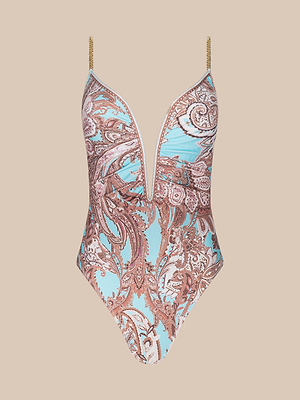 The Lilou One-Piece Swimsuit features a deep V neckline, blue background with a brown paisley pattern, thin brass-chain straps, and a high-leg cut. Shown against a beige backdrop.