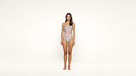 A woman stands barefoot against a plain white background, wearing a colorful, fitted one-piece swimsuit with a detailed pattern.