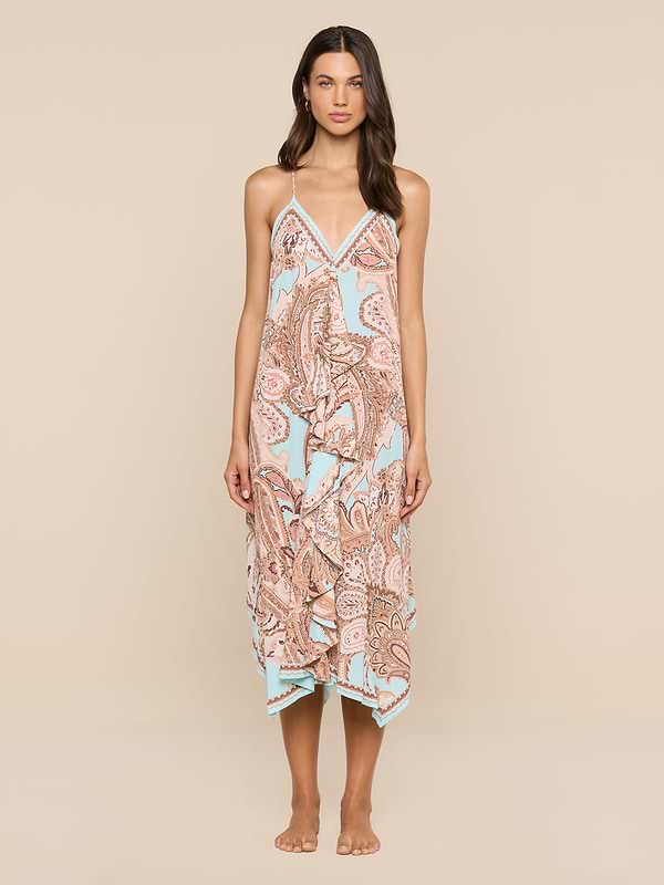 A woman stands barefoot against a white background, wearing the Elise Cover-Up Dress—a halter midi dress crafted from water-safe crepe. The dress features a paisley print in pastel colors, thin straps, and a handkerchief hem.