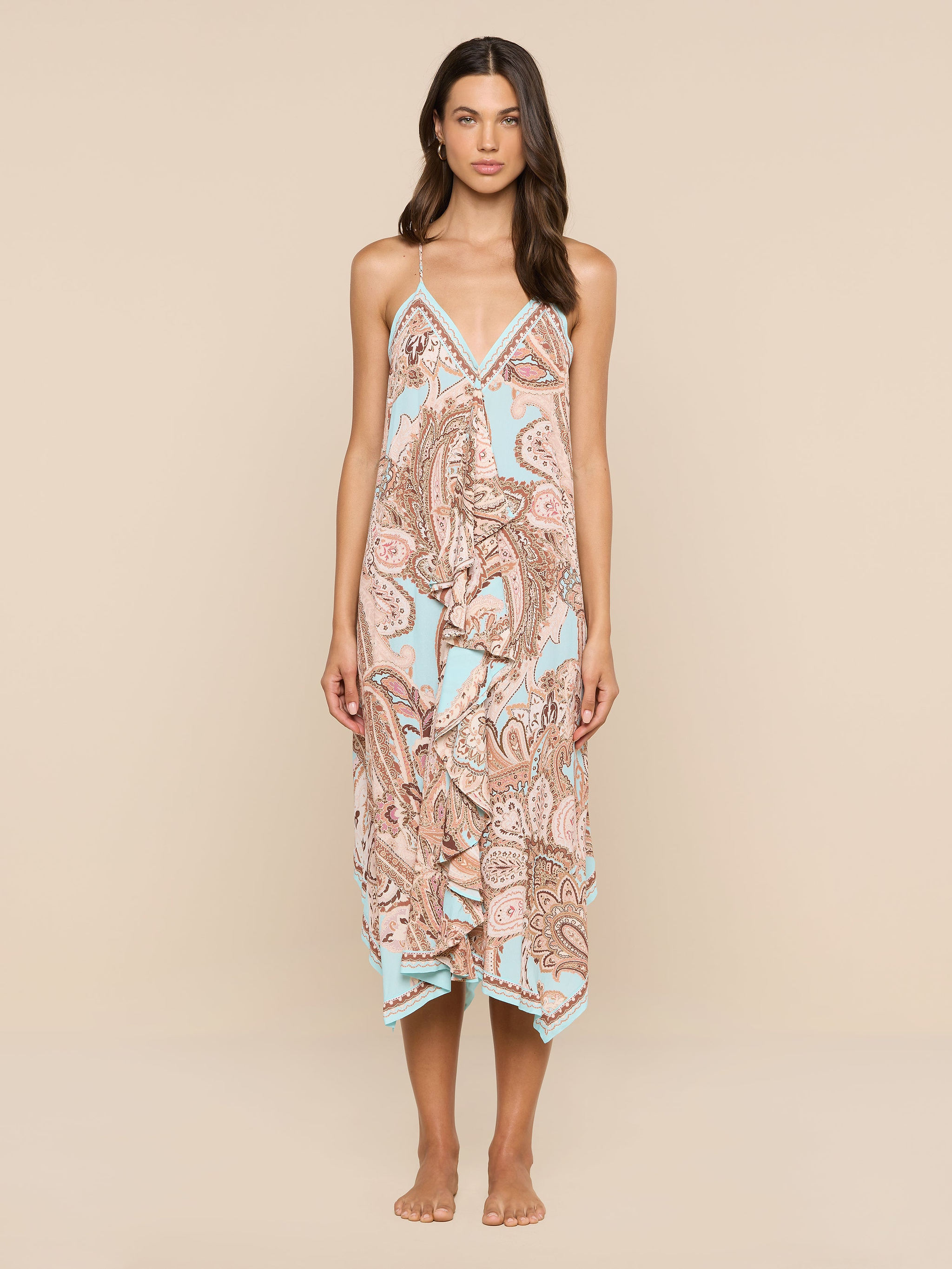 A woman stands barefoot against a white background, wearing the Elise Cover-Up Dress—a halter midi dress crafted from water-safe crepe. The dress features a paisley print in pastel colors, thin straps, and a handkerchief hem.