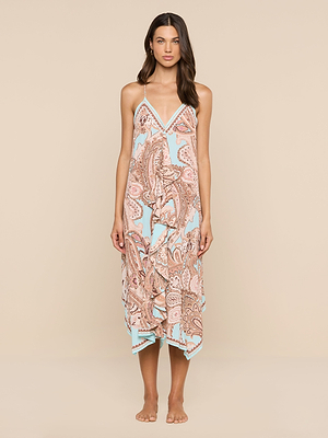 A woman with long, dark hair and a neutral expression stands barefoot against a beige background, wearing the Elise Cover-Up Dress in light blue and peach paisley print. This water-safe crepe dress features an asymmetrical hem, thin straps, and a sleeveless halter design.