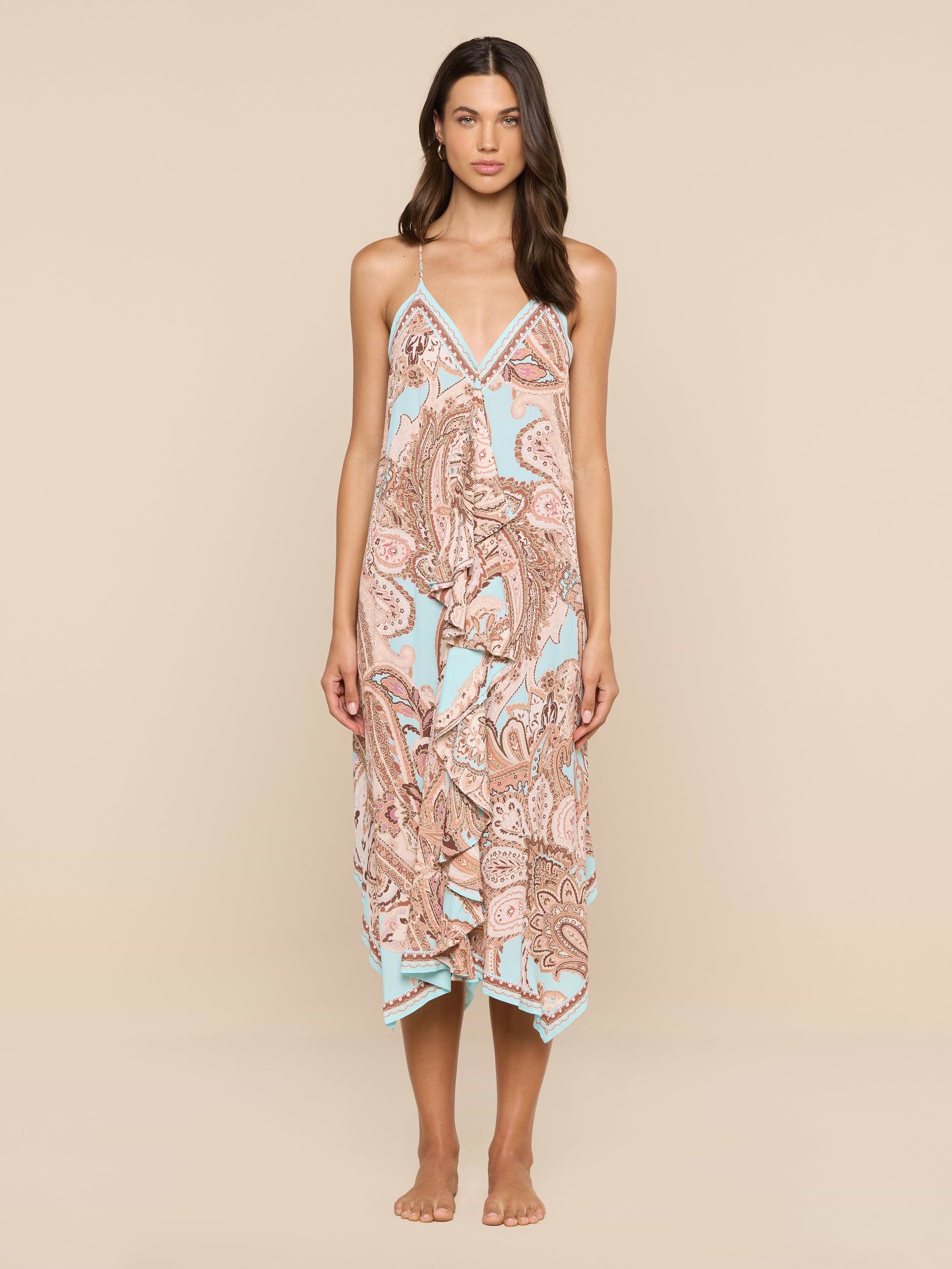 A woman stands barefoot against a white background, wearing the Elise Cover-Up Dress—a halter midi dress crafted from water-safe crepe. The dress features a paisley print in pastel colors, thin straps, and a handkerchief hem.