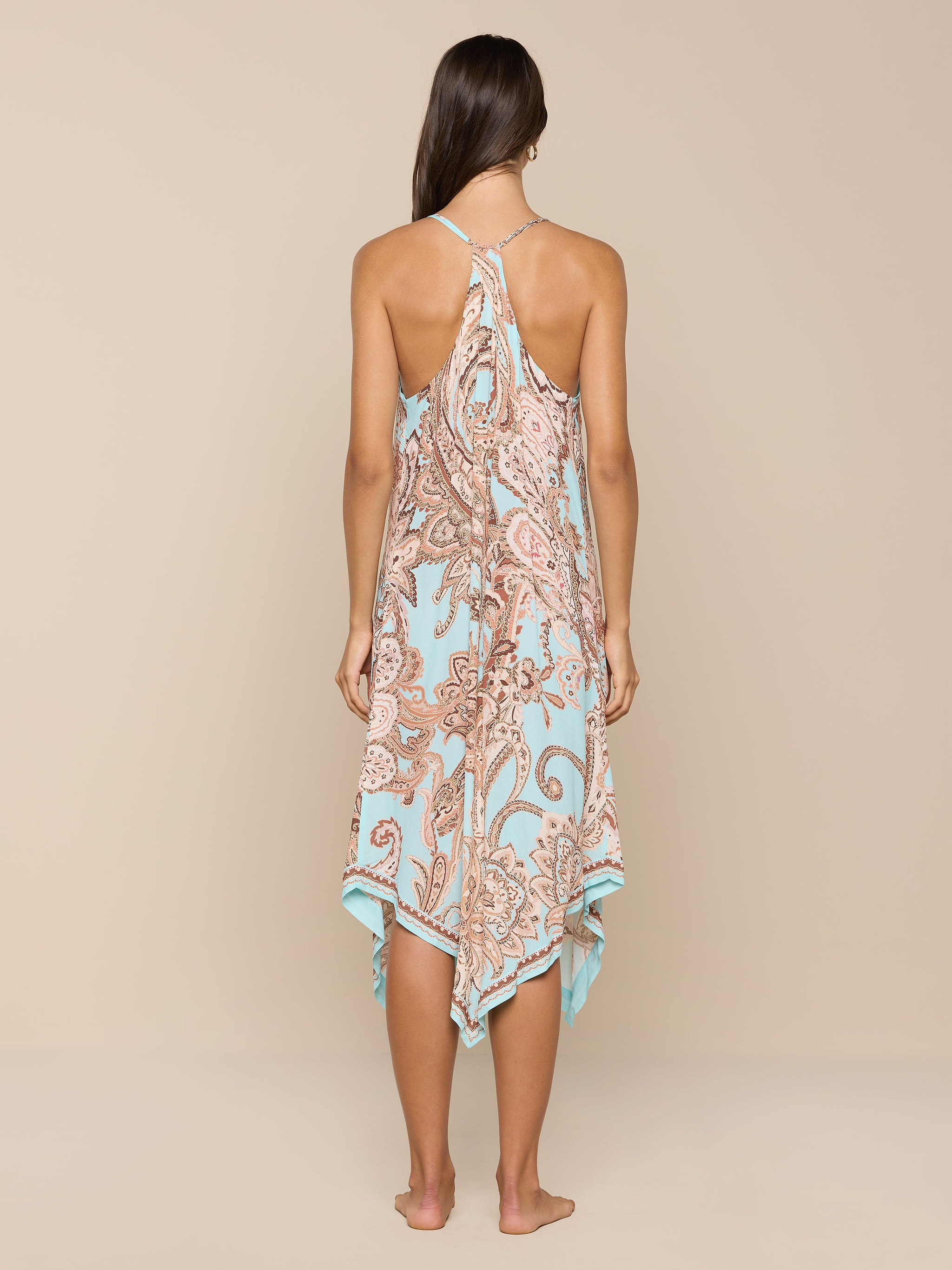 A woman stands barefoot, facing away, wearing the Elise Cover-Up Dress in a light blue and brown paisley print with a halter neckline and an uneven hem, set against a plain white background.