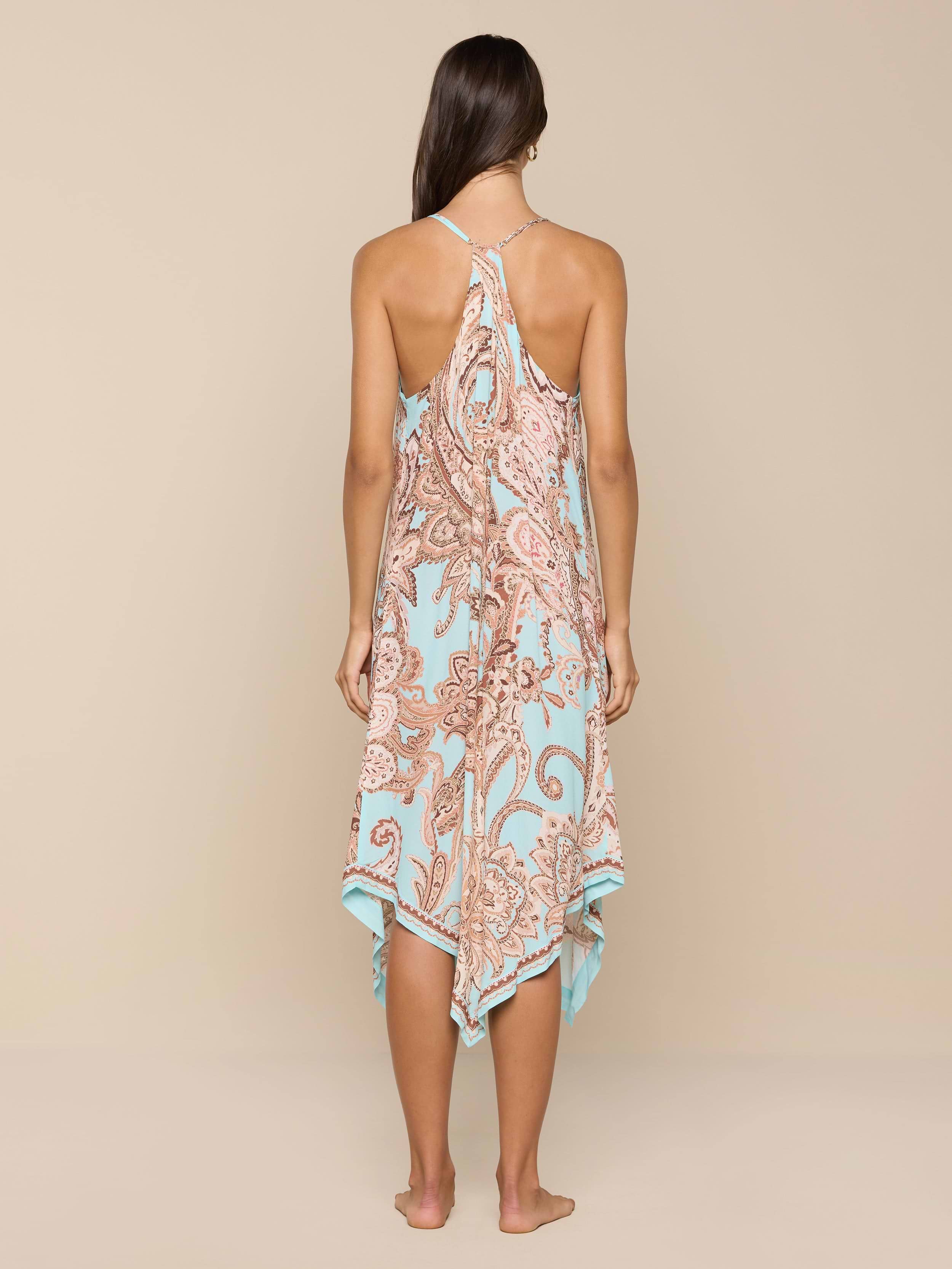 A woman stands barefoot, facing away, wearing the Elise Cover-Up Dress in a light blue and brown paisley print with a halter neckline and an uneven hem, set against a plain white background.