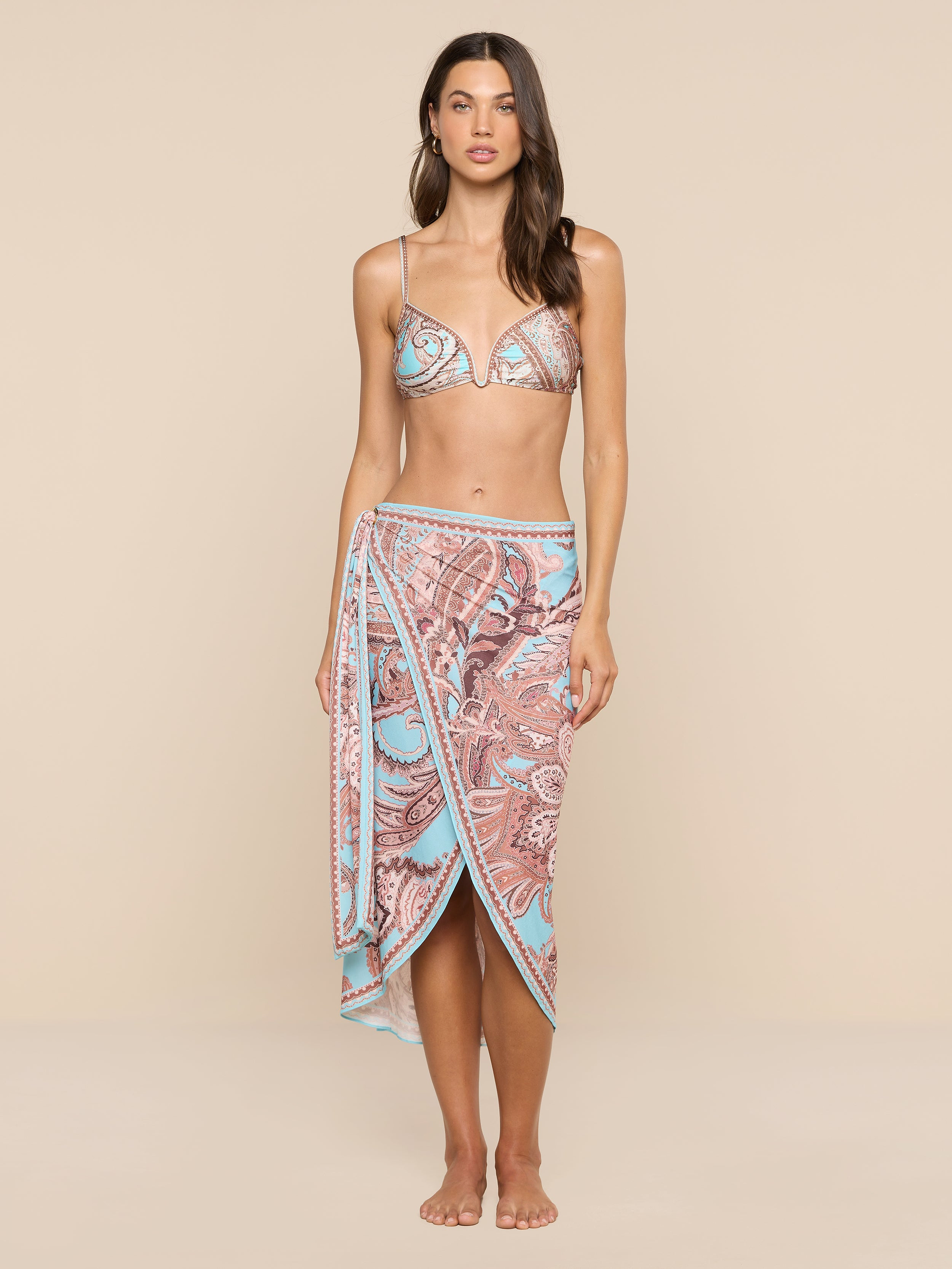 A woman stands barefoot against a plain white background, exuding beachside sophistication in a patterned bikini top and the Marena Sarong, which features matching blue and pink designs.