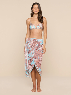 A woman with long brown hair stands barefoot against a beige background, wearing a patterned bikini top and the Marena Sarong—a blue and pink paisley skirt. She has a relaxed expression.