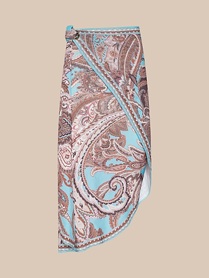 The Marena Sarong is a mid-length wrap skirt with a blue and brown paisley pattern, an asymmetrical hem, and a waistband buckle—ideal as a chic beach cover-up—shown against a beige background.