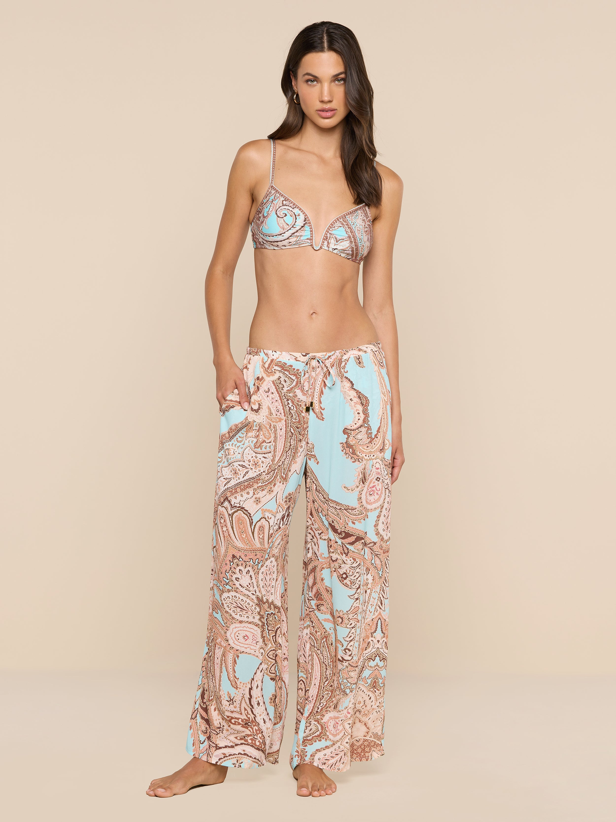 A woman stands barefoot in resort wear, featuring a paisley print bikini top paired with the Geraldine Cover-Up Pant—loose and relaxed in silhouette, showcasing light blue and brown tones—against a plain white background.