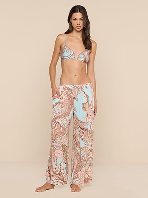 A woman poses against a beige background wearing the Geraldine Cover-Up Pant in a light blue and beige paisley print, with one hand in her pocket for a relaxed, resort-ready look.