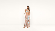 A woman stands barefoot in front of a plain white background, wearing a paisley bikini top and matching wide-leg pants in pastel blue and pink tones.