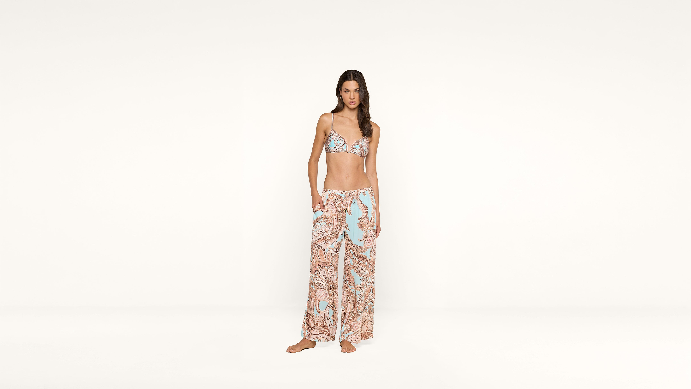 A woman stands barefoot in front of a plain white background, wearing a paisley bikini top and matching wide-leg pants in pastel blue and pink tones.