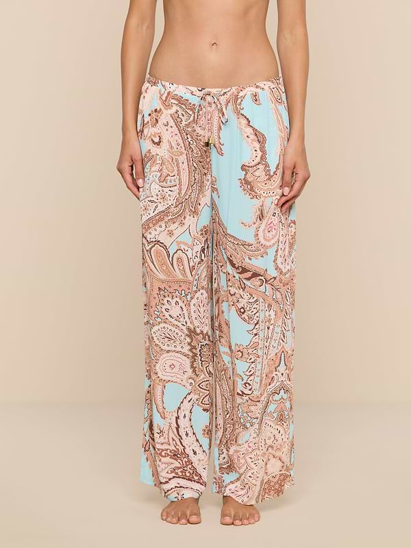 A person stands barefoot wearing the Geraldine Cover-Up Pant, featuring a wide-leg, light blue and tan paisley print with a relaxed silhouette. The image is cropped from the chest down, showcasing the pants against a plain white background.