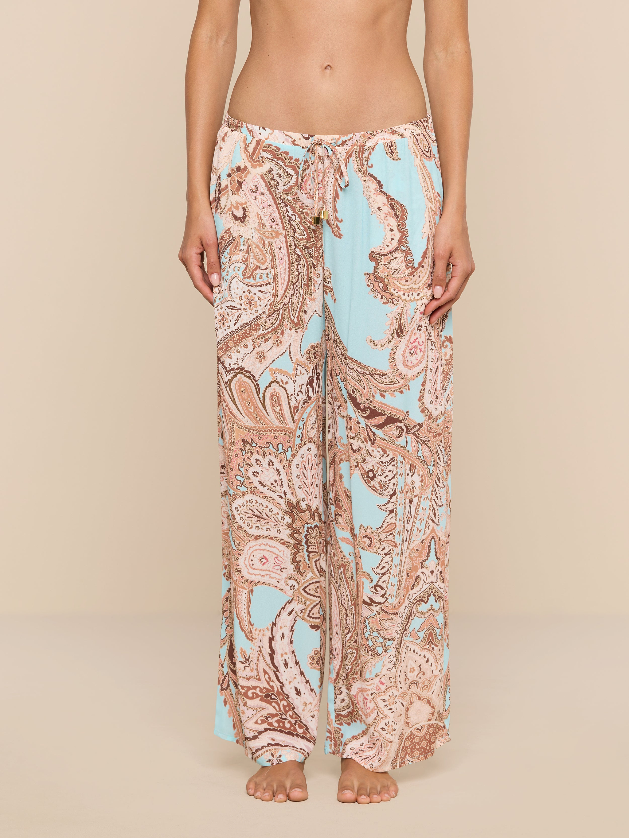 A person stands barefoot wearing the Geraldine Cover-Up Pant, featuring a wide-leg, light blue and tan paisley print with a relaxed silhouette. The image is cropped from the chest down, showcasing the pants against a plain white background.