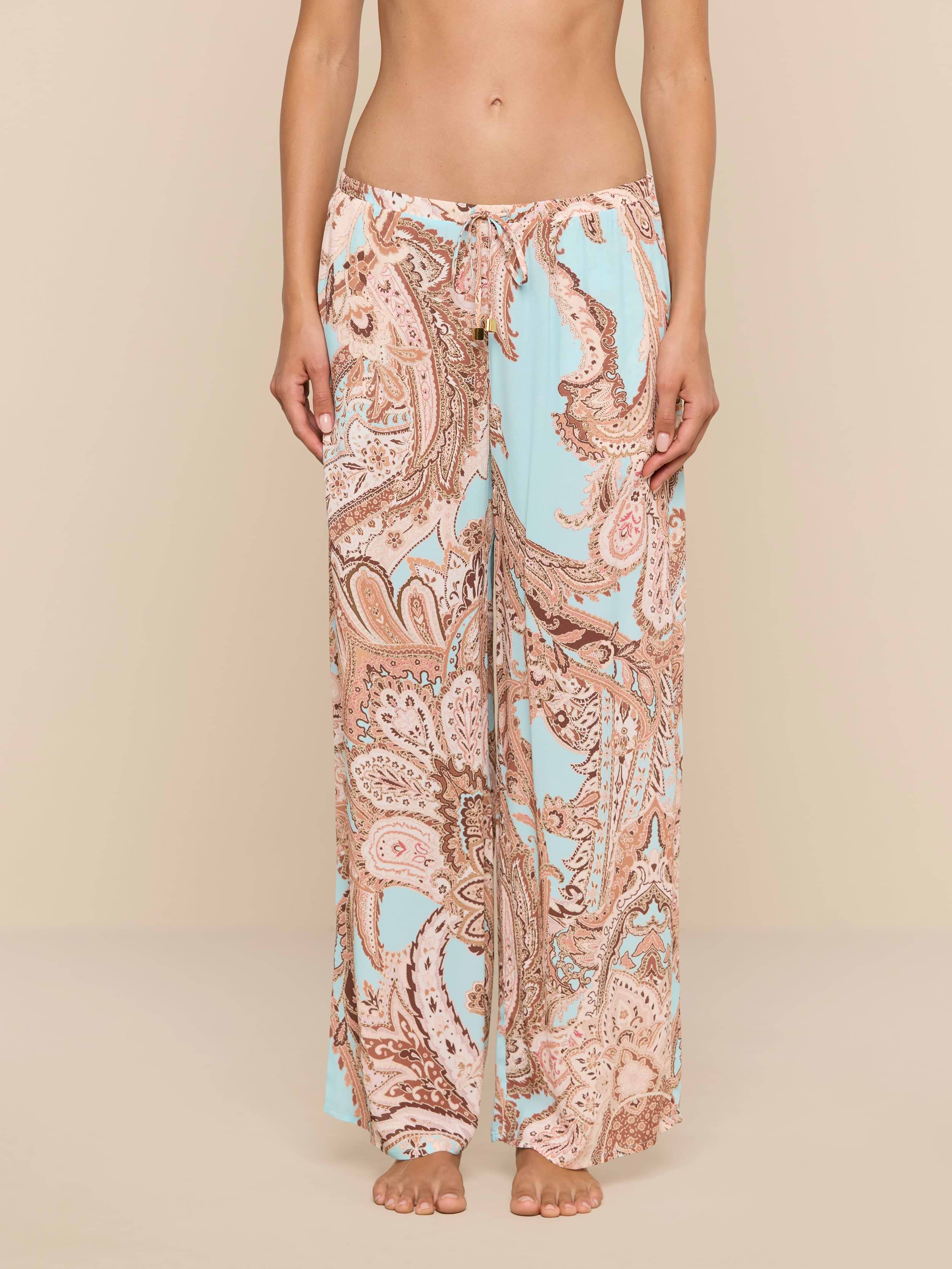 A person stands barefoot wearing the Geraldine Cover-Up Pant, featuring a wide-leg, light blue and tan paisley print with a relaxed silhouette. The image is cropped from the chest down, showcasing the pants against a plain white background.
