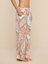 A person stands barefoot in side profile, wearing the Geraldine Cover-Up Pant—a high-waisted, loose-fitting style adorned with a pastel paisley print in pink and light blue hues. With its relaxed silhouette, this stylish piece embodies the effortless elegance of resort wear against a plain white background.