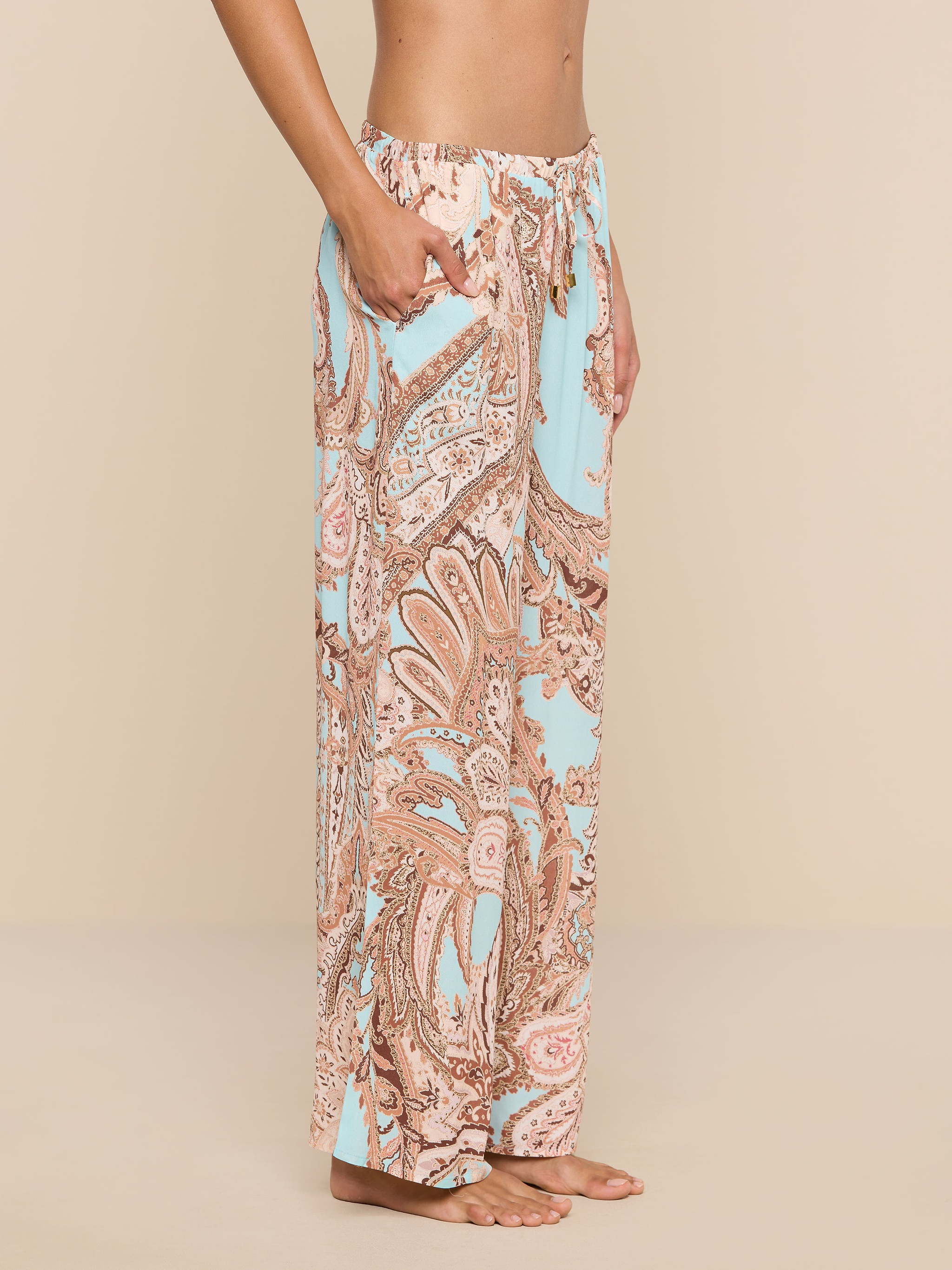 A person stands barefoot in side profile, wearing the Geraldine Cover-Up Pant—a high-waisted, loose-fitting style adorned with a pastel paisley print in pink and light blue hues. With its relaxed silhouette, this stylish piece embodies the effortless elegance of resort wear against a plain white background.