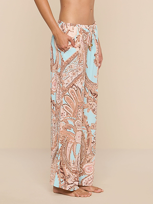 A person stands barefoot on a neutral background, wearing the Geraldine Cover-Up Pant—light blue and beige paisley-print wide-leg pants with a relaxed silhouette, elastic drawstring waist, and side pockets. Only the lower half is visible.