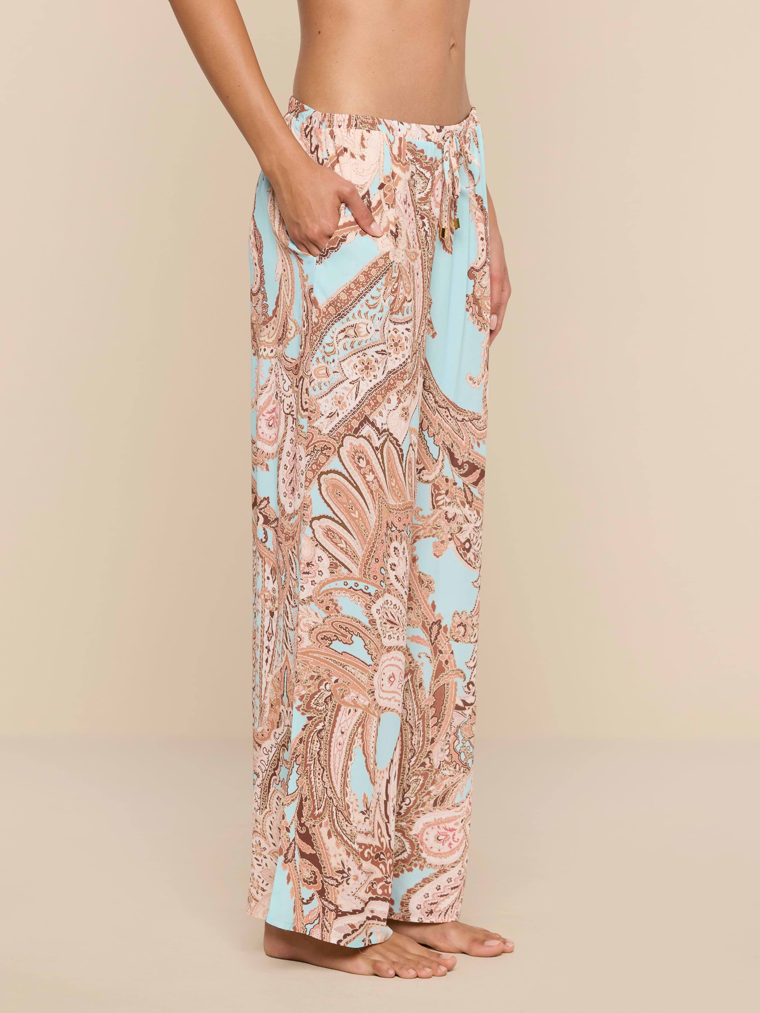 A person stands barefoot in side profile, wearing the Geraldine Cover-Up Pant—a high-waisted, loose-fitting style adorned with a pastel paisley print in pink and light blue hues. With its relaxed silhouette, this stylish piece embodies the effortless elegance of resort wear against a plain white background.