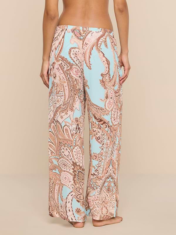 A person stands barefoot, facing away, wearing the Geraldine Cover-Up Pant—a loose-fitting, wide-leg style featuring a light blue and tan paisley print. The relaxed silhouette of the pants adds to the effortless feel against the plain white background.