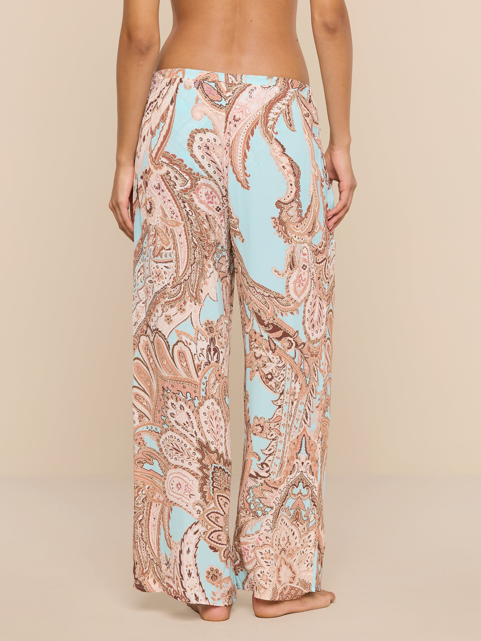 A person stands barefoot, facing away, wearing the Geraldine Cover-Up Pant—a loose-fitting, wide-leg style featuring a light blue and tan paisley print. The relaxed silhouette of the pants adds to the effortless feel against the plain white background.