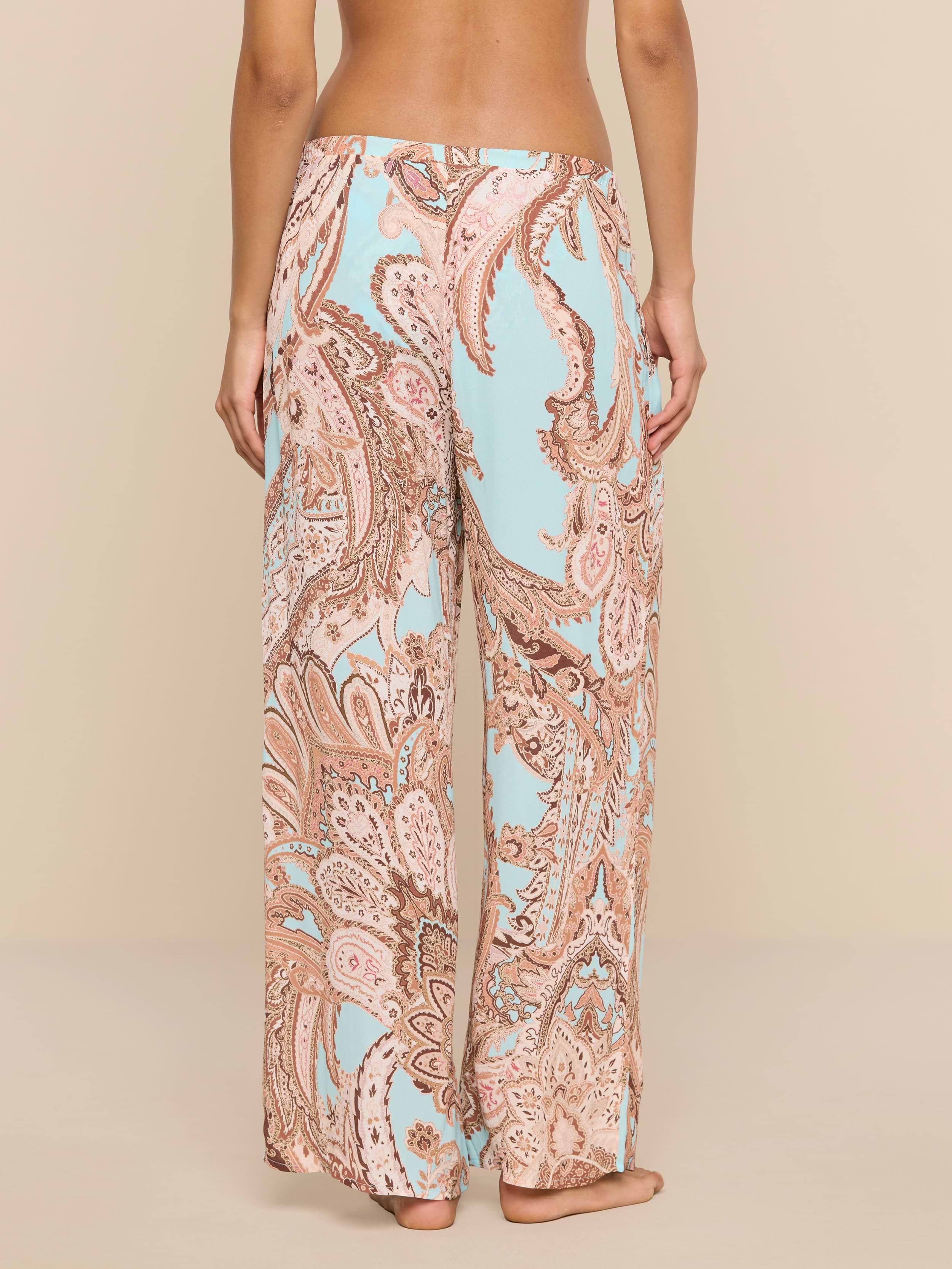 A person stands barefoot, facing away, wearing the Geraldine Cover-Up Pant—a loose-fitting, wide-leg style featuring a light blue and tan paisley print. The relaxed silhouette of the pants adds to the effortless feel against the plain white background.